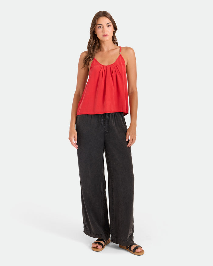 A woman with long brown hair faces forward against a white background, wearing Volcom's Women's Washed Up Twill Pants—wide-leg, tie-front style with an elastic waist—paired with a loose red sleeveless top and black sandals.