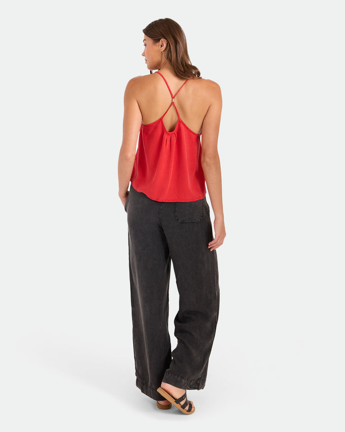 A woman stands facing away against a white background, wearing Volcom Women's Washed Up Twill Pants with an elastic waist, paired with a red strappy top and sandals. Her hair is down.